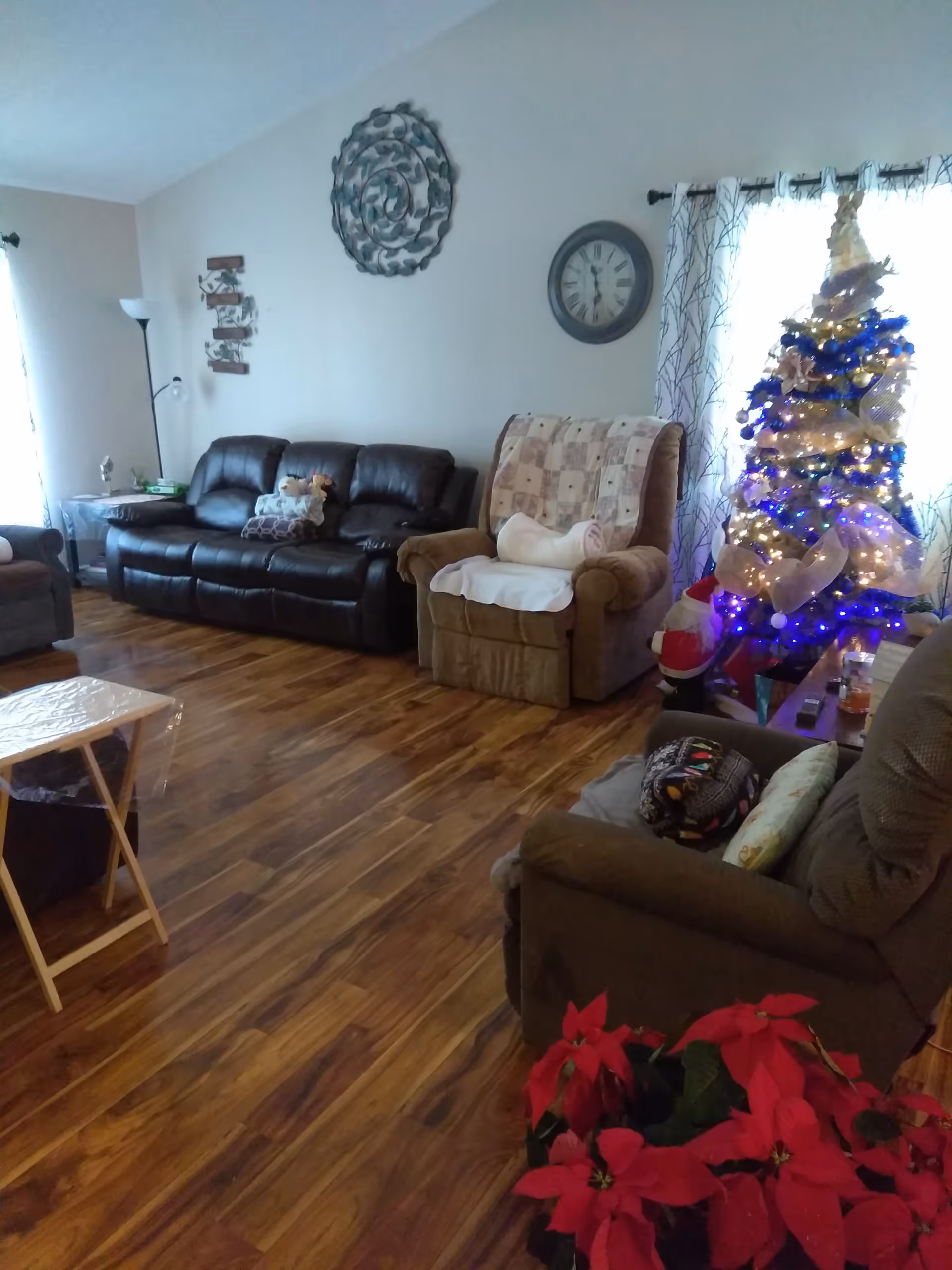 A cozy living room with wooden flooring, featuring a black leather sofa, a brown recliner with a quilt and pillows, another cushioned chair, a decorated Christmas tree with lights, and a poinsettia plant in the foreground. The walls have a decorative metal wall art and a round clock. Light filters in through windows with patterned curtains.