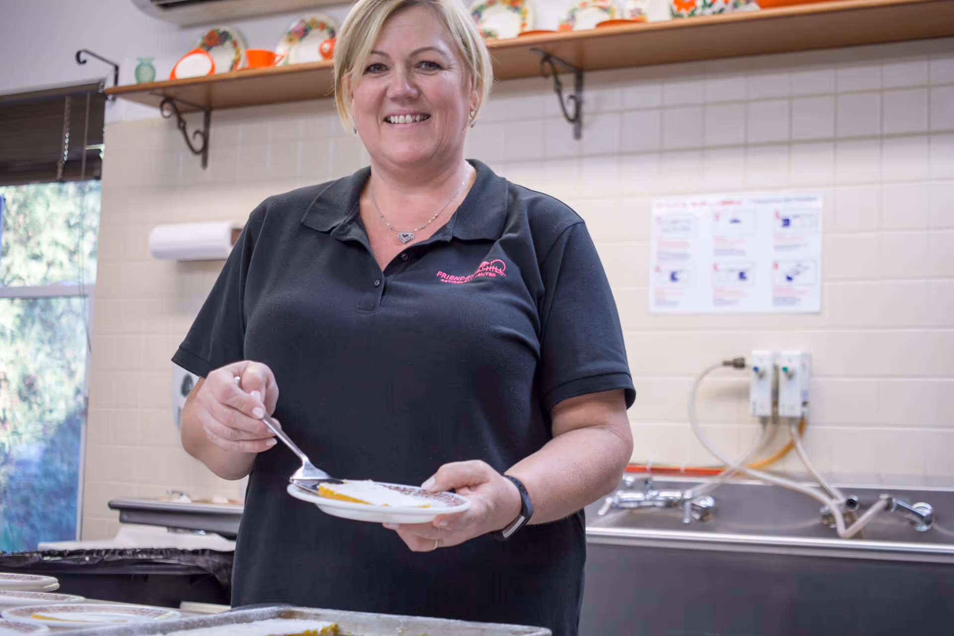 A staff member in a black polo scoops food onto a plate in a tiled kitchen with shelves of dishes.