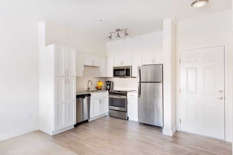 A modern kitchen with white cabinets, stainless steel appliances including a refrigerator, microwave, dishwasher, and oven. There is a sink with a faucet, a coffee maker on the counter, and a bowl of fruit. The floor is light wood, and there is a white door to the right.