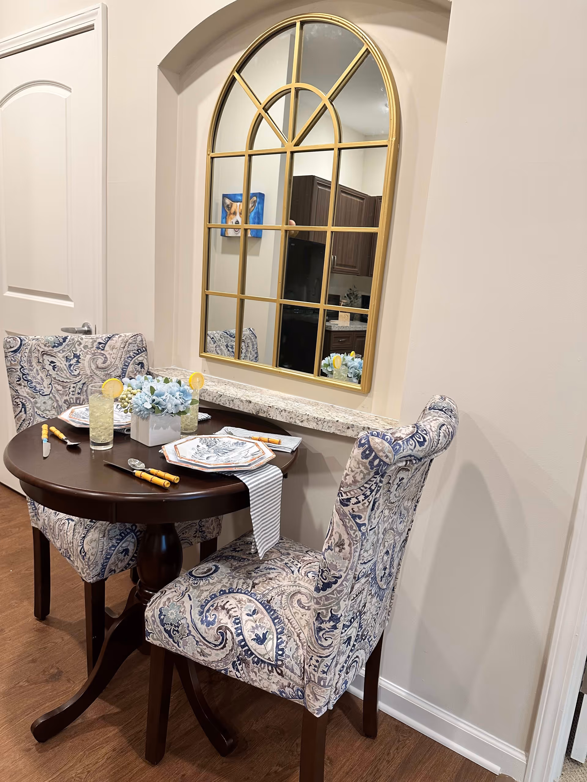 A cozy dining table setting featuring two upholstered chairs, a round table with plates and utensils, and a decorative mirror in the background.