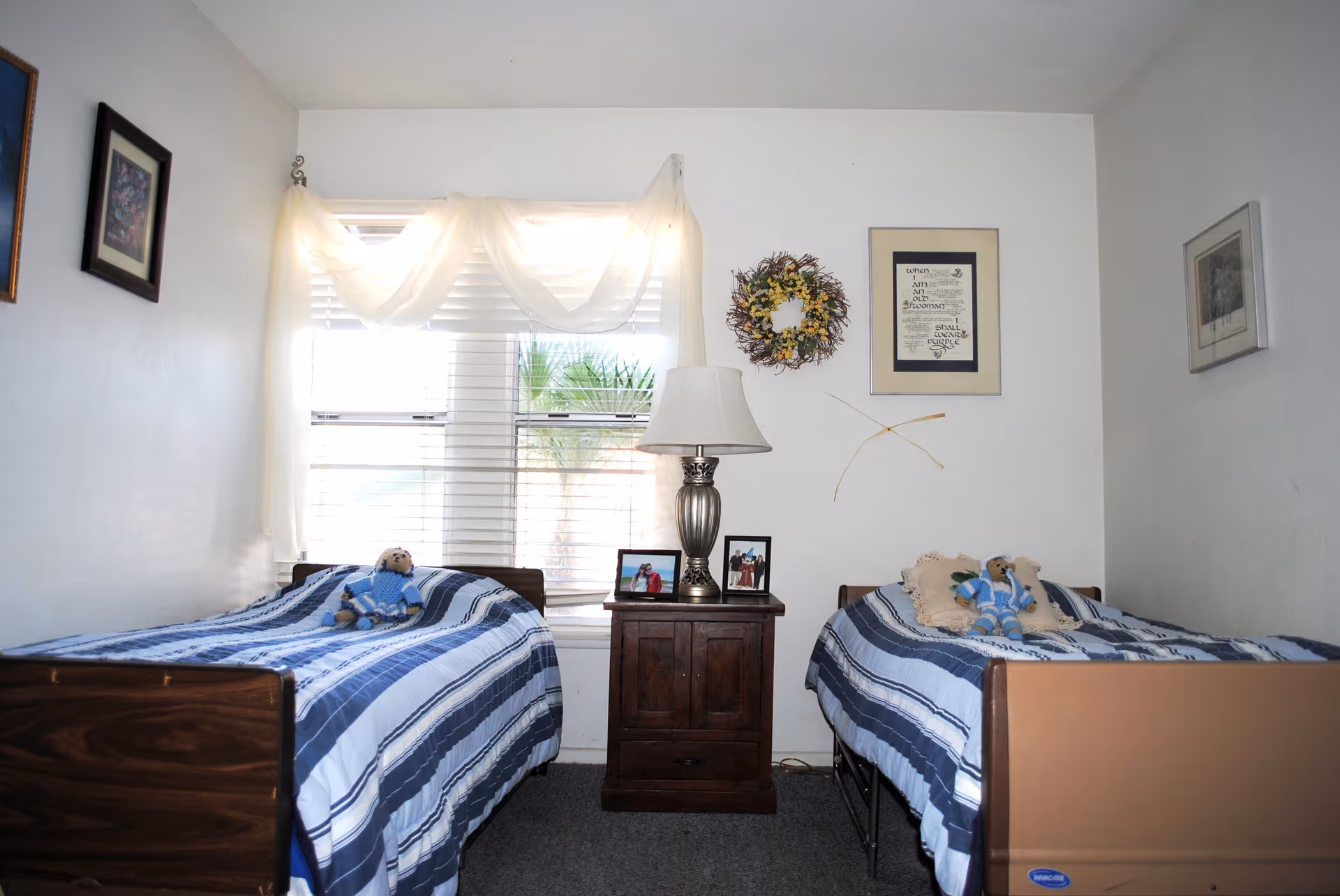 A small bedroom with two single beds on either side of a wooden nightstand. Each bed has a blue and white striped bedspread and a teddy bear dressed in blue sitting on top. The nightstand holds a silver lamp and two framed photographs. A window with white blinds and sheer curtains is behind the nightstand. The walls are decorated with framed pictures and a floral wreath.