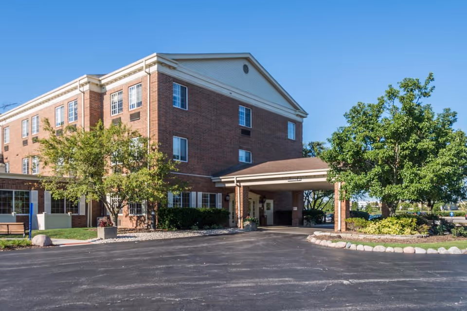 Exterior view of Avantara Libertyville, a three-story brick building with multiple windows, a covered entrance, and surrounding greenery including trees and shrubs under a clear blue sky.
