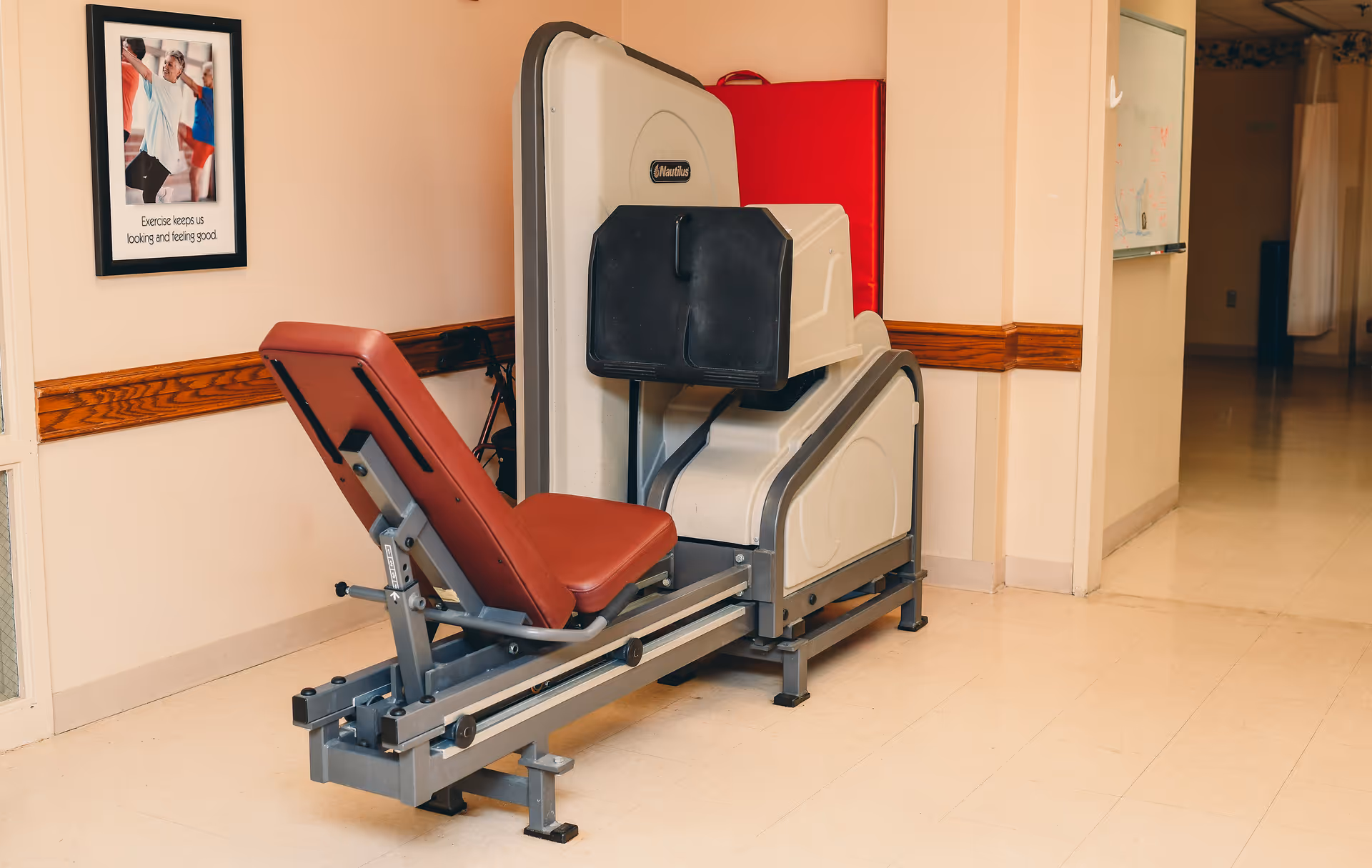 Exercise machine with a red padded seat and backrest in a hallway with beige walls and a framed poster on the wall showing people exercising with the text 'Exercise keeps us looking and feeling good.'
