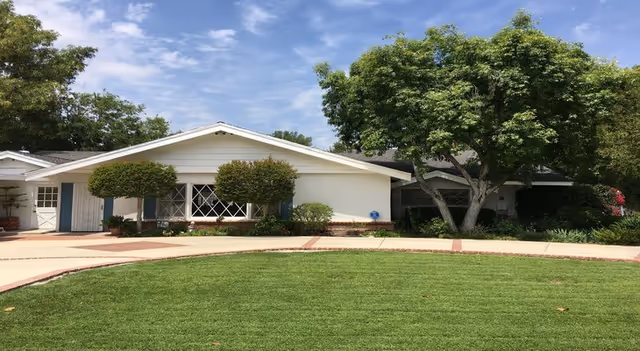 Single-story residential building with white exterior walls and a sloped roof, surrounded by neatly trimmed bushes and trees, with a well-maintained green lawn in front under a partly cloudy blue sky.
