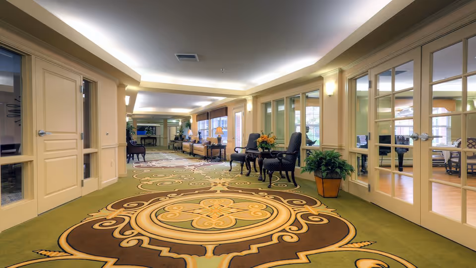 A spacious and well-lit hallway in a senior living community with a decorative carpet featuring intricate patterns. The hallway has beige walls, multiple doors with glass panels on the right side, and several chairs and small tables with flower arrangements along the walls. Large windows allow natural light to enter, creating a welcoming atmosphere.