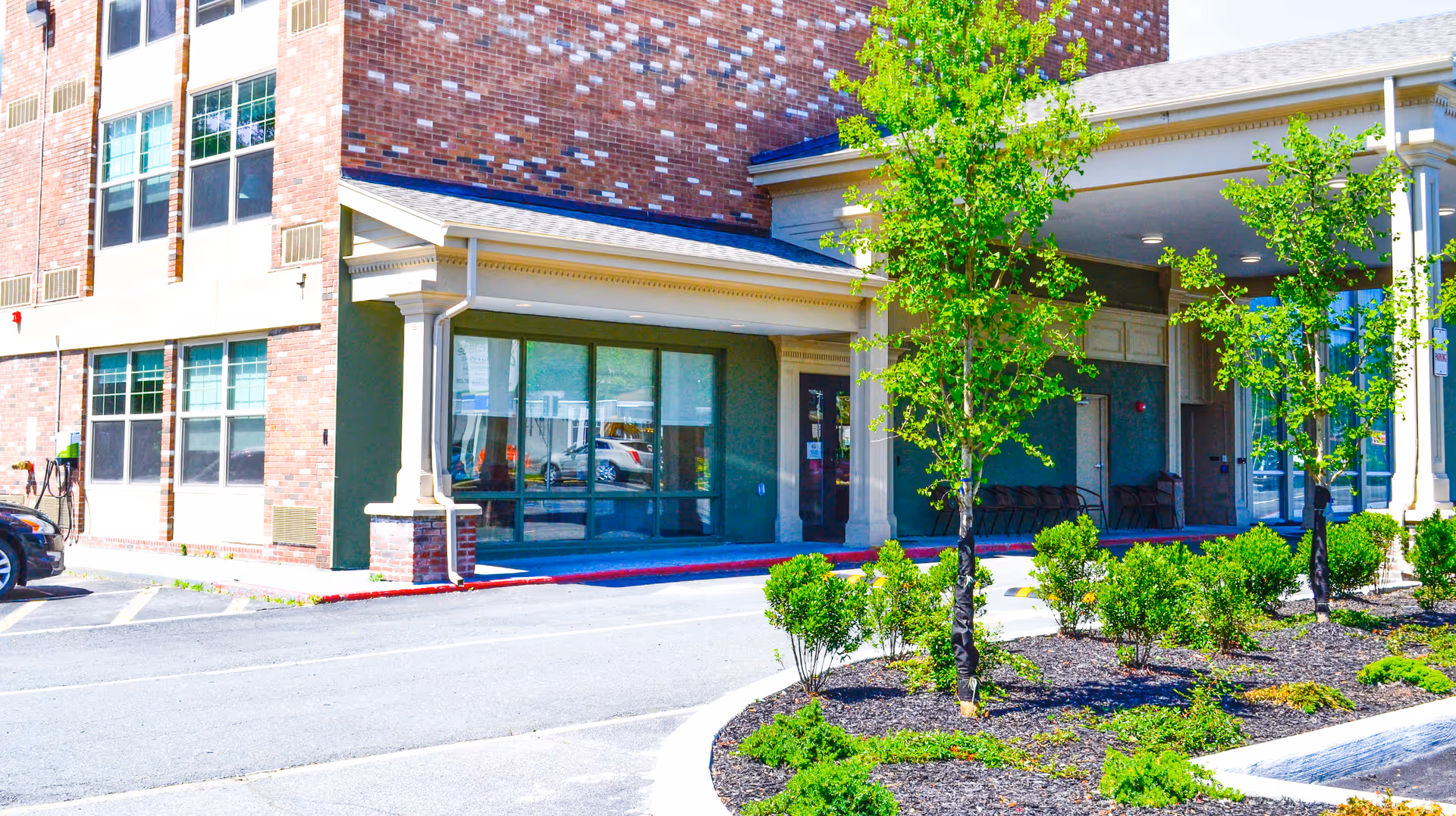 Entrance area of a senior living facility named Promenade at University Place, showing a brick building with large windows, a covered drop-off area, small trees, and landscaped bushes in front.