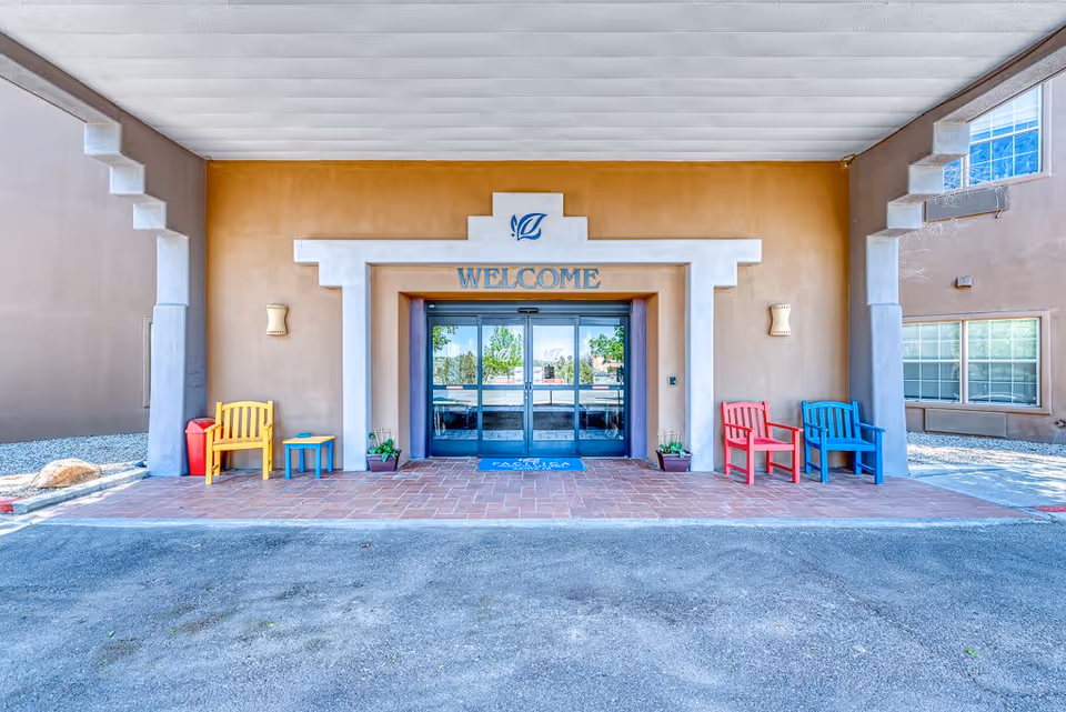 Entrance to a building with a large 'WELCOME' sign above automatic glass doors. The entrance area has a tiled floor and is flanked by colorful chairs—yellow and red on the left side, red and blue on the right side—along with small tables and potted plants. The building exterior is beige with white architectural accents.