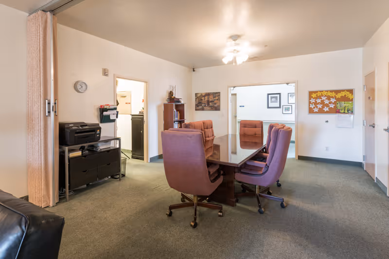 A meeting or conference room with a rectangular wooden table surrounded by six purple office chairs on wheels. The room has beige walls and green carpet. There is a black printer on a metal stand with drawers, a clock on the wall, a bulletin board with star-shaped notes, and framed pictures on the walls. Two doorways lead to other rooms.