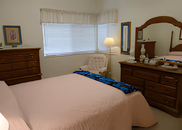 A bedroom with a large bed covered in a light pink bedspread, a wooden dresser with a mirror, a wooden chest of drawers with a framed picture on top, a white cushioned chair, a floor lamp, and a window with sheer curtains.