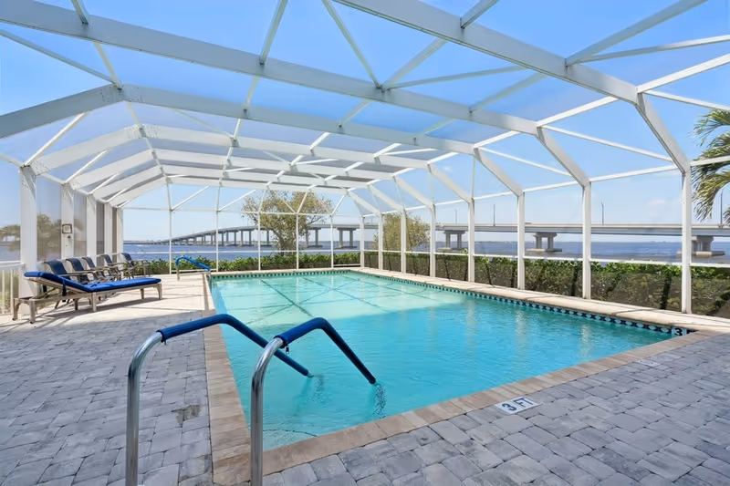 Indoor swimming pool with clear blue water enclosed by a white screened structure. There are lounge chairs with blue cushions on the left side and a view of a bridge and water body in the background under a clear blue sky.