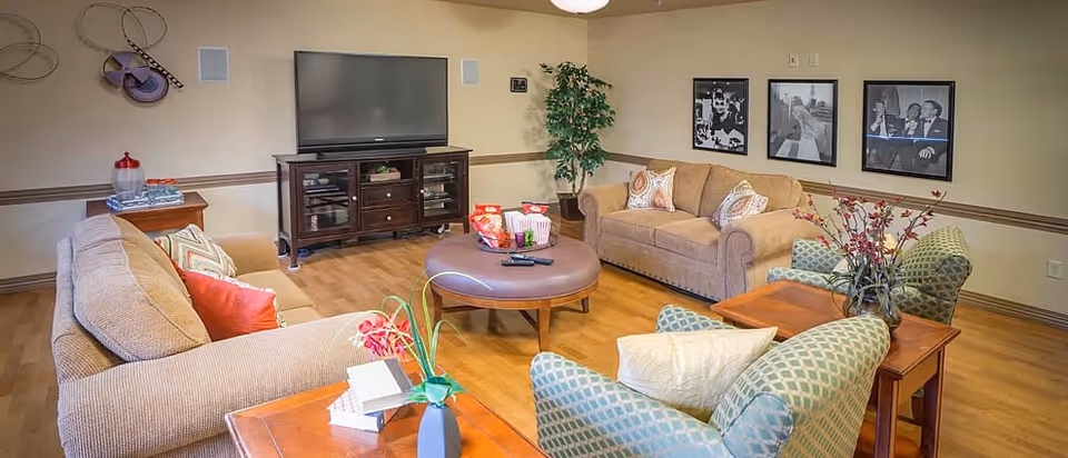 A cozy living room with two beige sofas, two patterned armchairs, a round coffee table with snacks and remote controls, a wooden TV stand with a flat-screen television, wall art, and a potted plant in the corner.