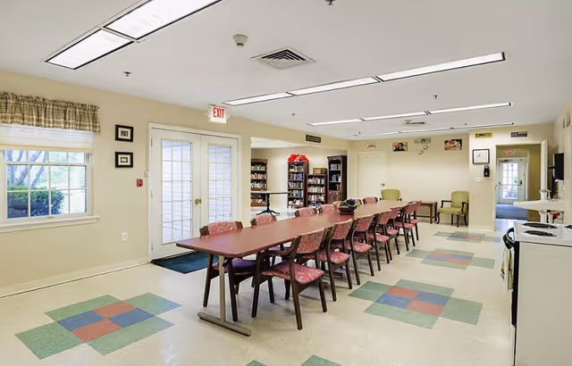 A spacious common area in a senior living facility featuring a long table with multiple chairs, a bookshelf filled with books, two armchairs, and a kitchenette area with a stove. The room has large windows, double glass doors, and colorful square patterns on the floor tiles.