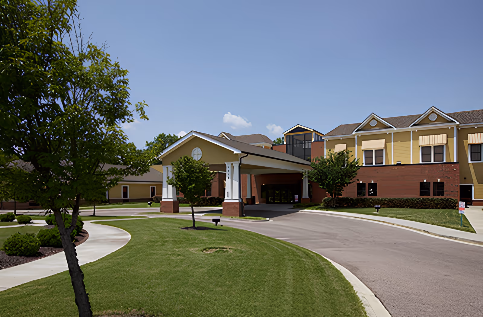 Exterior view of AHC Savannah senior living facility showing a driveway leading to a covered entrance with yellow and brick buildings in the background, surrounded by green grass and trees under a clear blue sky.