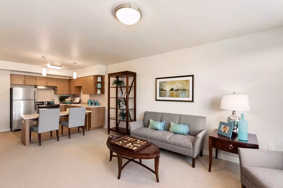 A modern open-concept living area and kitchen in a senior living facility. The living area features a gray sofa with three blue-green pillows, a wooden coffee table with a backgammon game, a side table with a lamp, a framed photo, and decorative items. The kitchen has light wood cabinets, stainless steel refrigerator and stove, a small island with two gray upholstered chairs, and pendant lights hanging above.