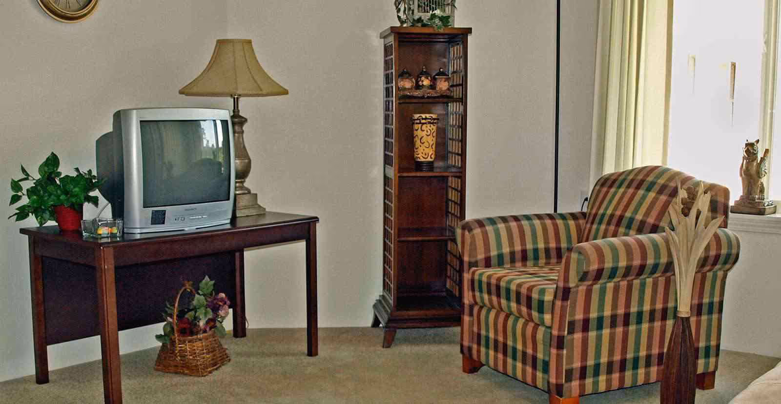 A cozy living room corner with a plaid armchair, a wooden side table holding a vintage TV and a lamp, a tall wooden shelving unit with decorative items, a basket with flowers on the floor, and a window with light curtains letting in natural light.