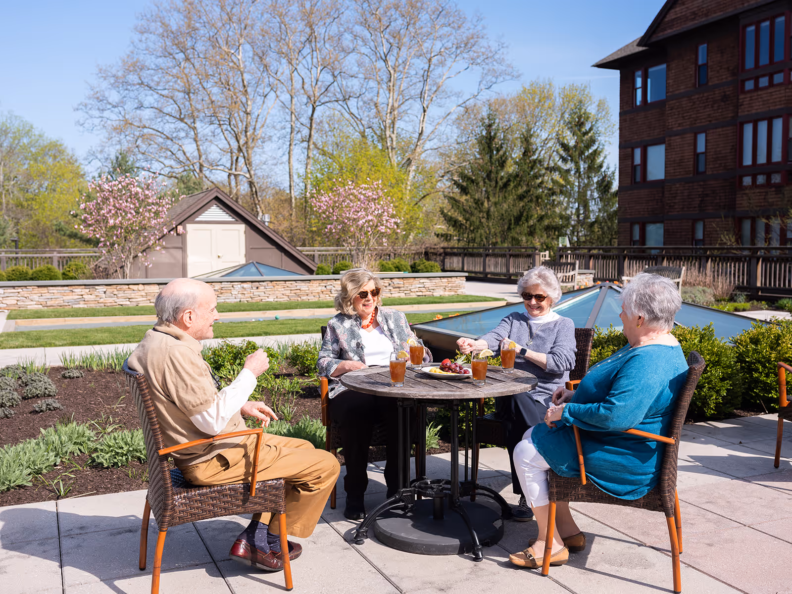 Four elderly people sitting around a round outdoor table on a sunny day, enjoying drinks and snacks, with trees and a building in the background.