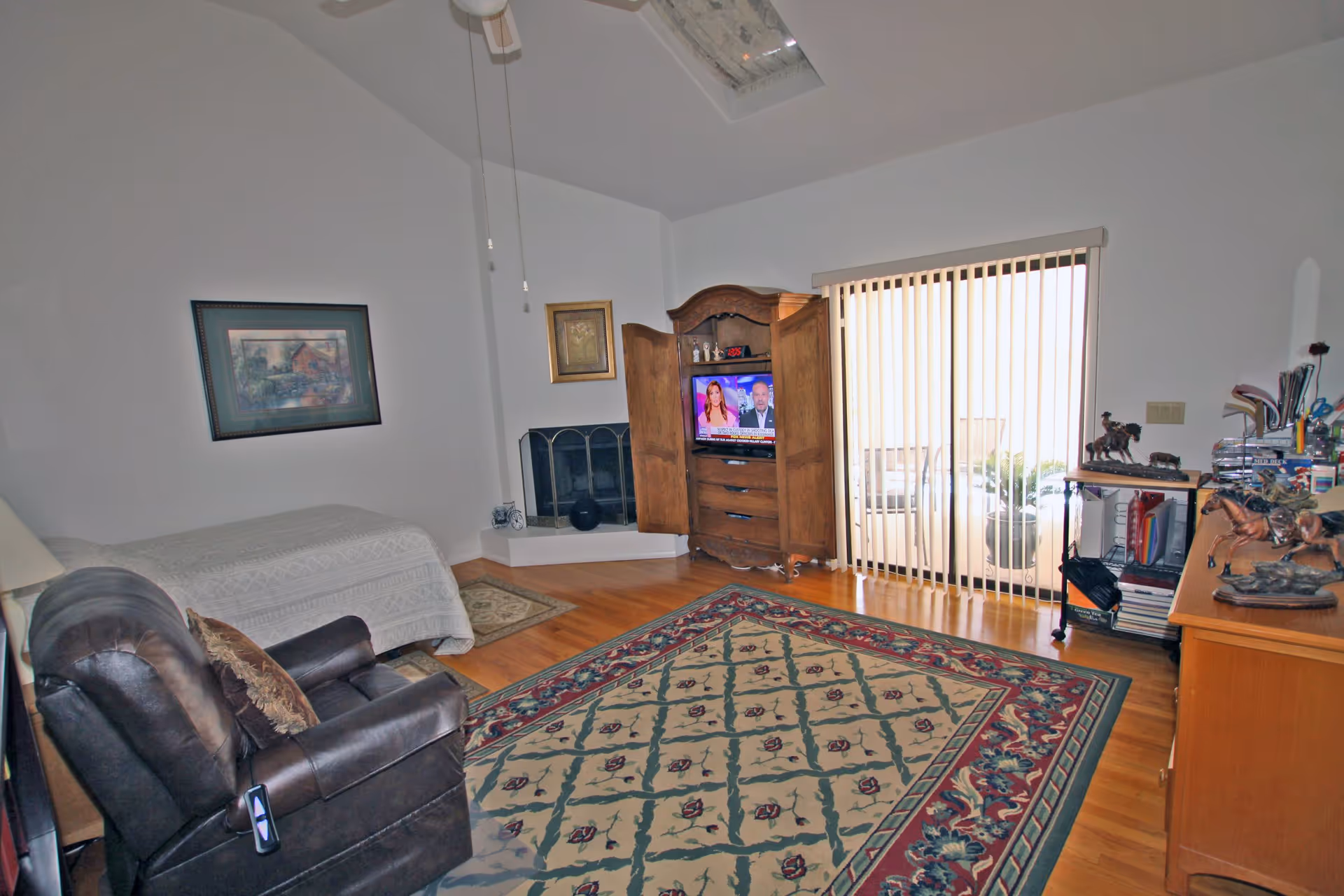 Spacious living room with a patterned area rug, leather recliner, wooden armoire with a TV, sliding glass door with vertical blinds, and a bed against the wall.