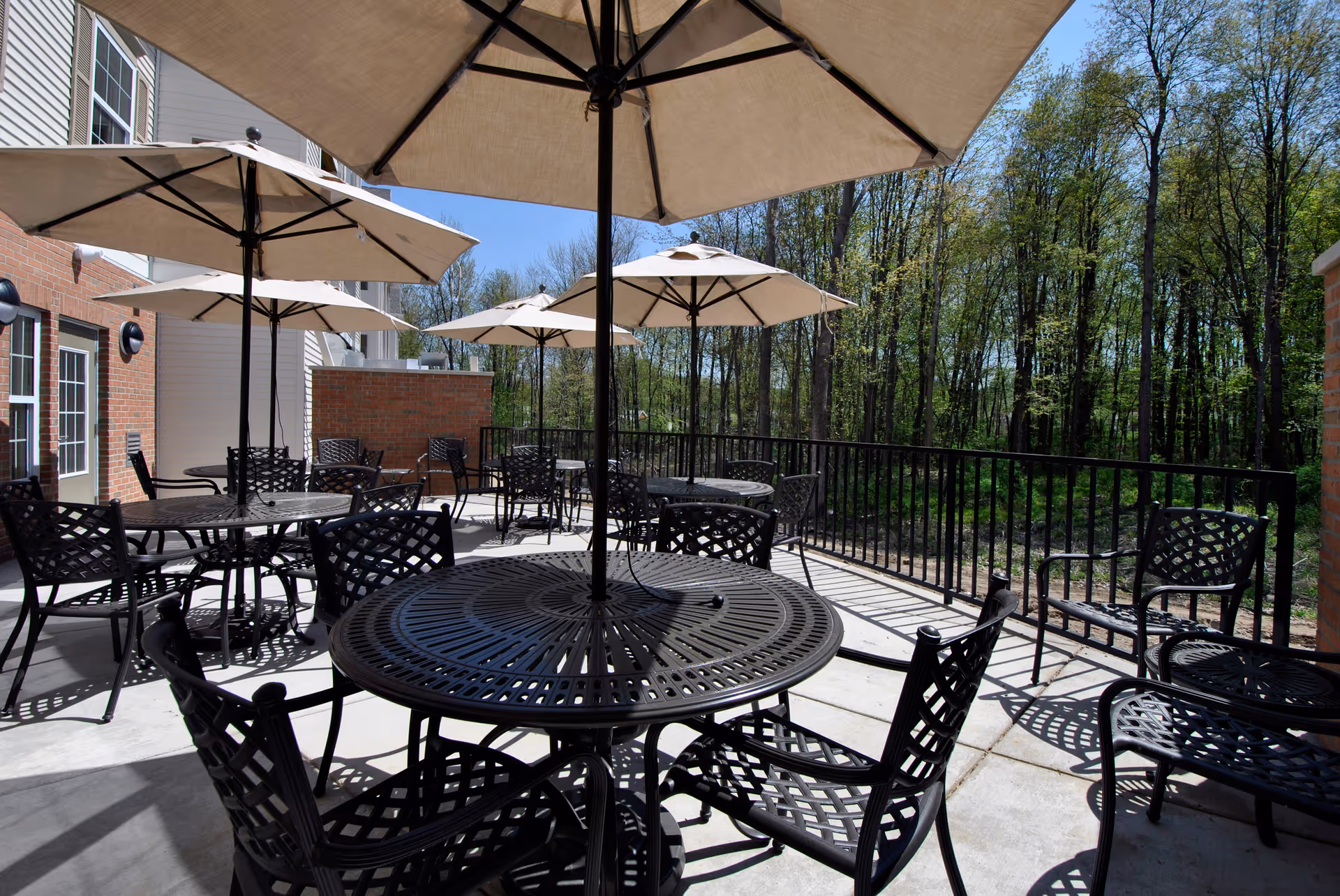 Outdoor patio area with multiple round metal tables and chairs, each table shaded by a large beige umbrella. The patio is adjacent to a brick and siding building, with a black metal railing along the edge. Trees and greenery are visible in the background under a clear blue sky.