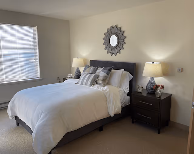 A neatly made bed with white bedding and multiple pillows in a bedroom. There are two dark wooden nightstands on either side of the bed, each with a lamp. A decorative round mirror with a floral frame hangs above the bed. A window with blinds is on the left wall, allowing natural light into the room.