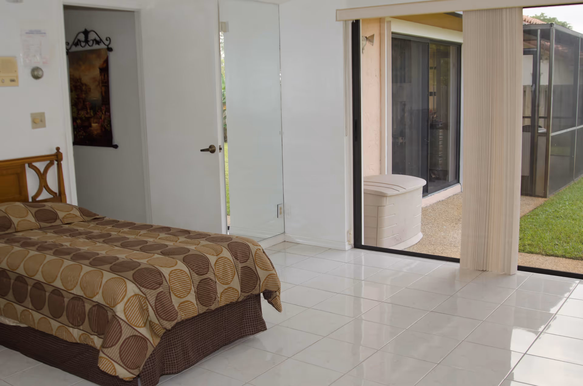 A bedroom with a bed covered in a brown and beige patterned bedspread. The room has white tiled floors, a white door slightly ajar, a large mirror on the wall, and a sliding glass door leading to an outdoor area with grass and a patio.