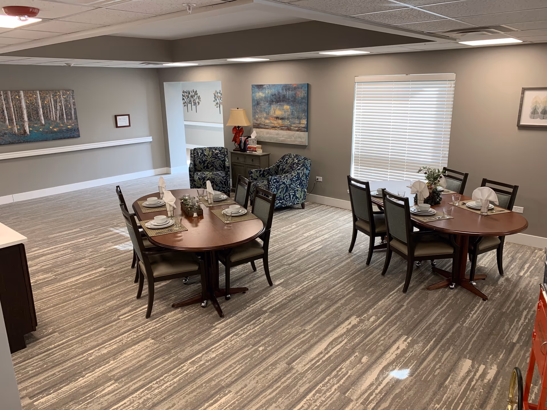 A dining area in a senior living facility with two wooden dining tables set with plates, bowls, glasses, and napkins. There are six chairs around each table. In the background, there are two patterned armchairs, a small cabinet with a lamp and books, and artwork on the walls. A window with closed blinds is also visible.