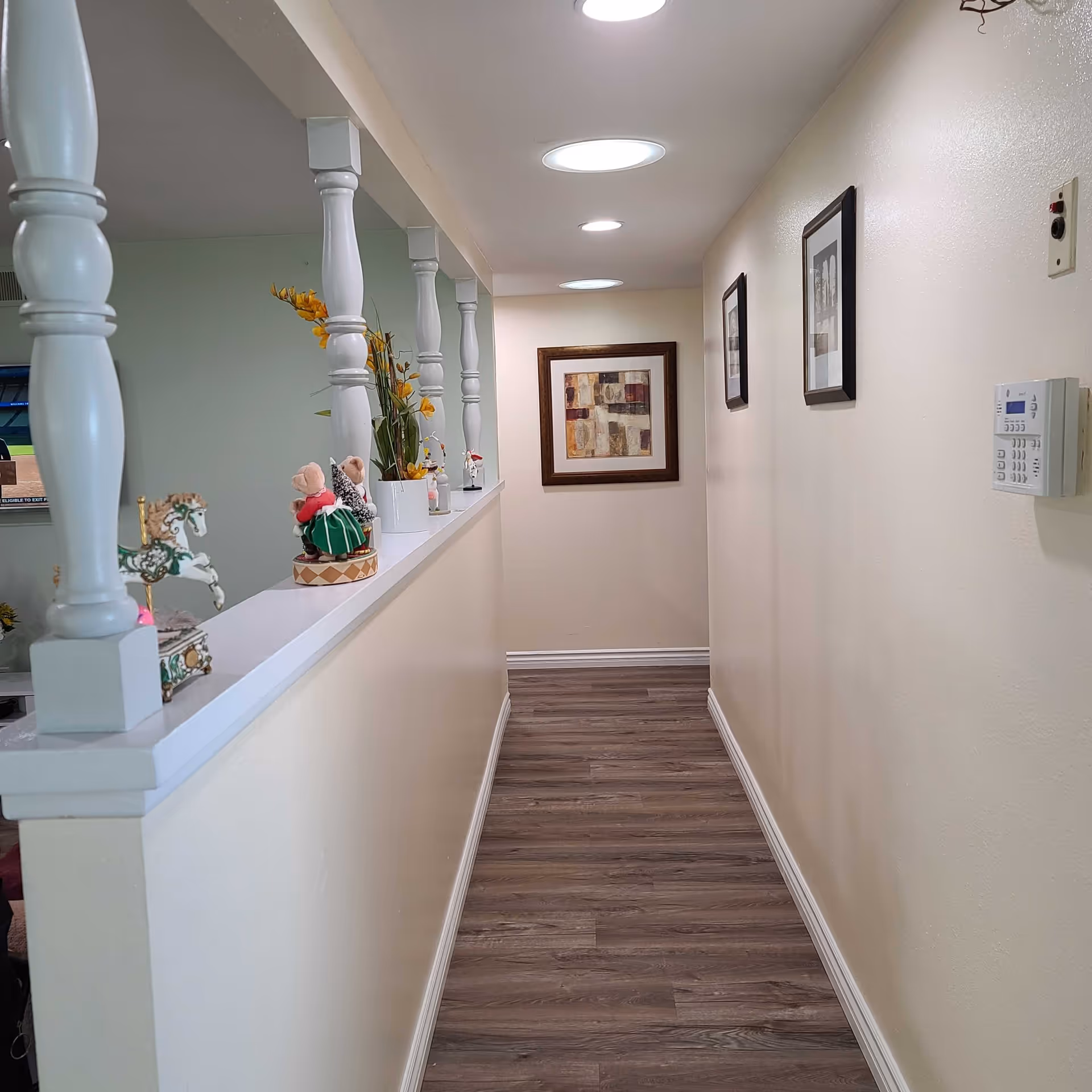 A narrow hallway with light-colored walls and wood flooring. On the right wall, there are three framed pictures and a security keypad. On the left side, there is a half wall with white decorative columns and various small decorative items placed on top. The hallway is lit by recessed ceiling lights.