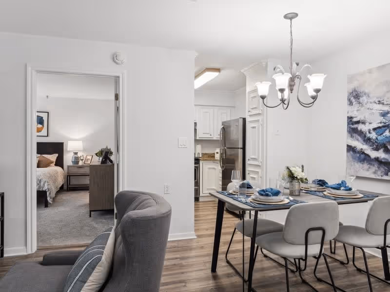 View of a modern apartment interior showing a dining area with a table set for four, a kitchen with white cabinets and stainless steel appliances, and a bedroom visible through an open doorway. The dining table has blue napkins and plates, and a chandelier hangs above it. The flooring is wood, and there is a large abstract painting on the wall.