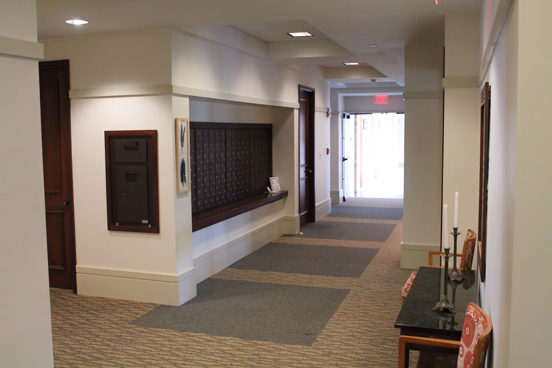 Interior hallway of The Summit at Brighton Independent Living with a row of mailboxes on the left wall, a small table with two chairs and candle holders on the right, and a door at the end of the hallway letting in natural light.