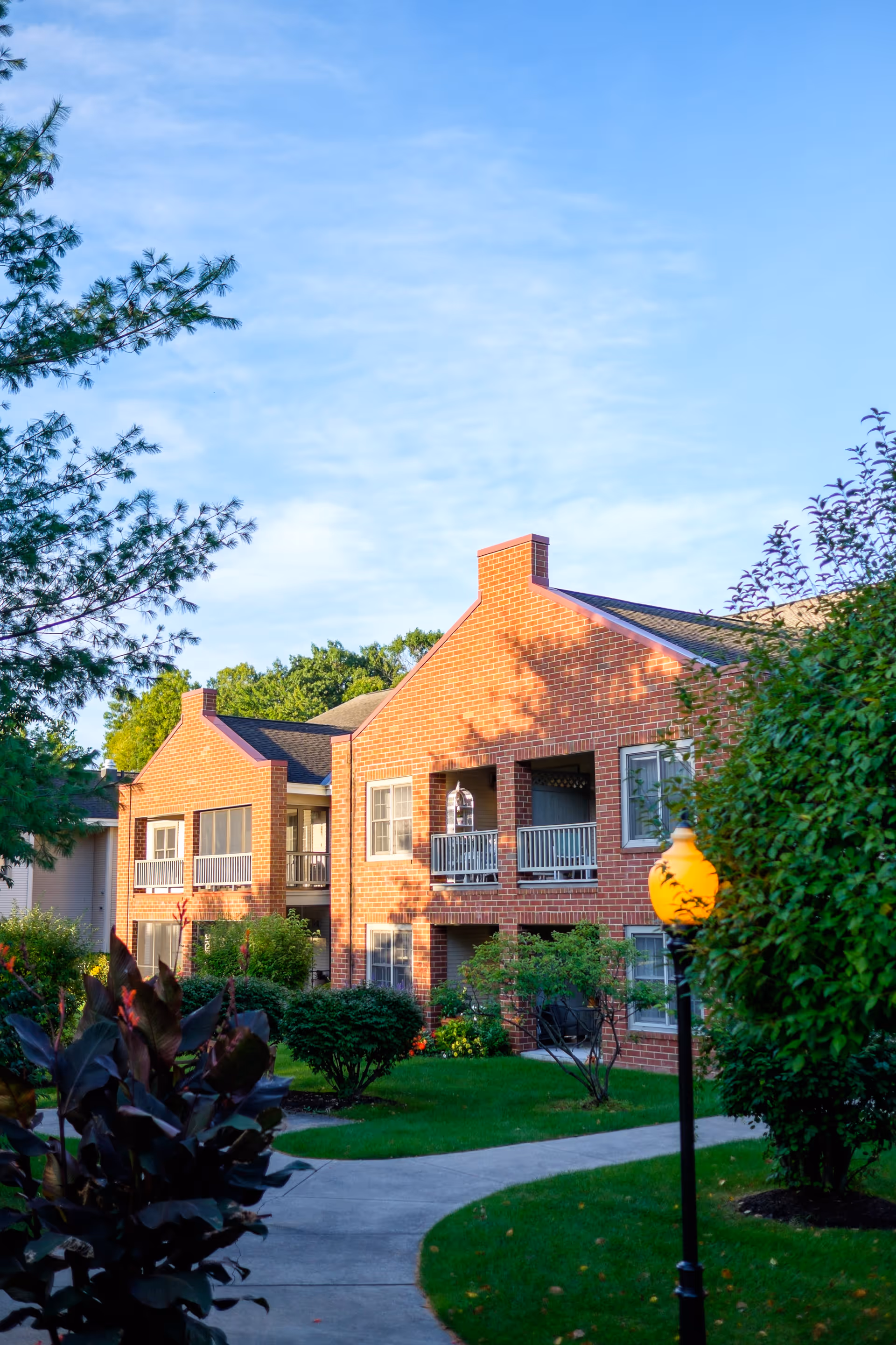 Exterior view of a two-story brick building with balconies, surrounded by green trees, bushes, and a well-maintained lawn under a clear blue sky. A curved concrete pathway and a lamp post with a yellow light are visible in the foreground.
