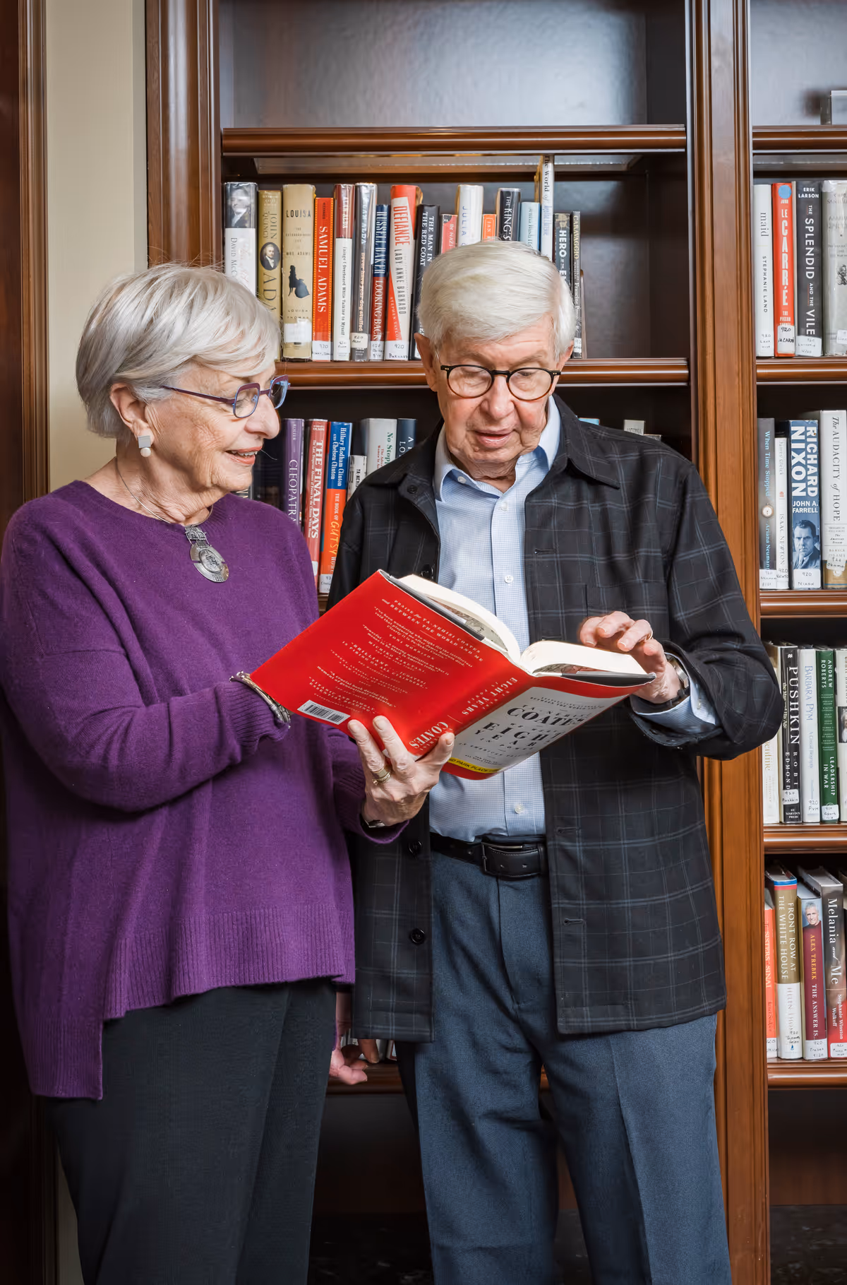 An elderly woman and an elderly man standing in front of a bookshelf filled with books. The woman, wearing a purple sweater and glasses, is holding a red book open while the man, wearing a dark jacket and glasses, looks at the book. They appear to be engaged in reading or discussing the book together.