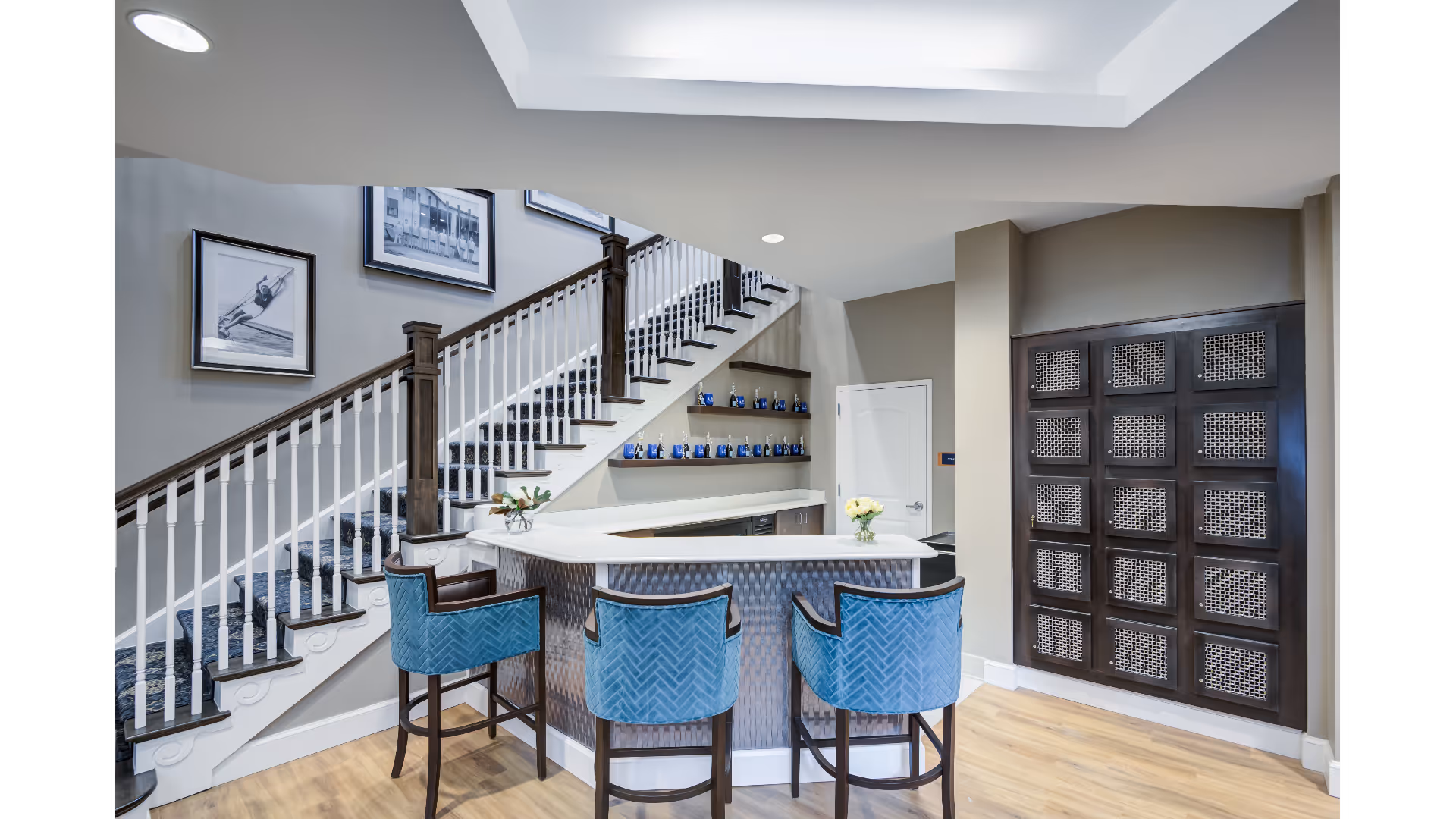 Interior lounge with a curved white bar, three blue barstools, and a staircase with framed artwork along the wall.