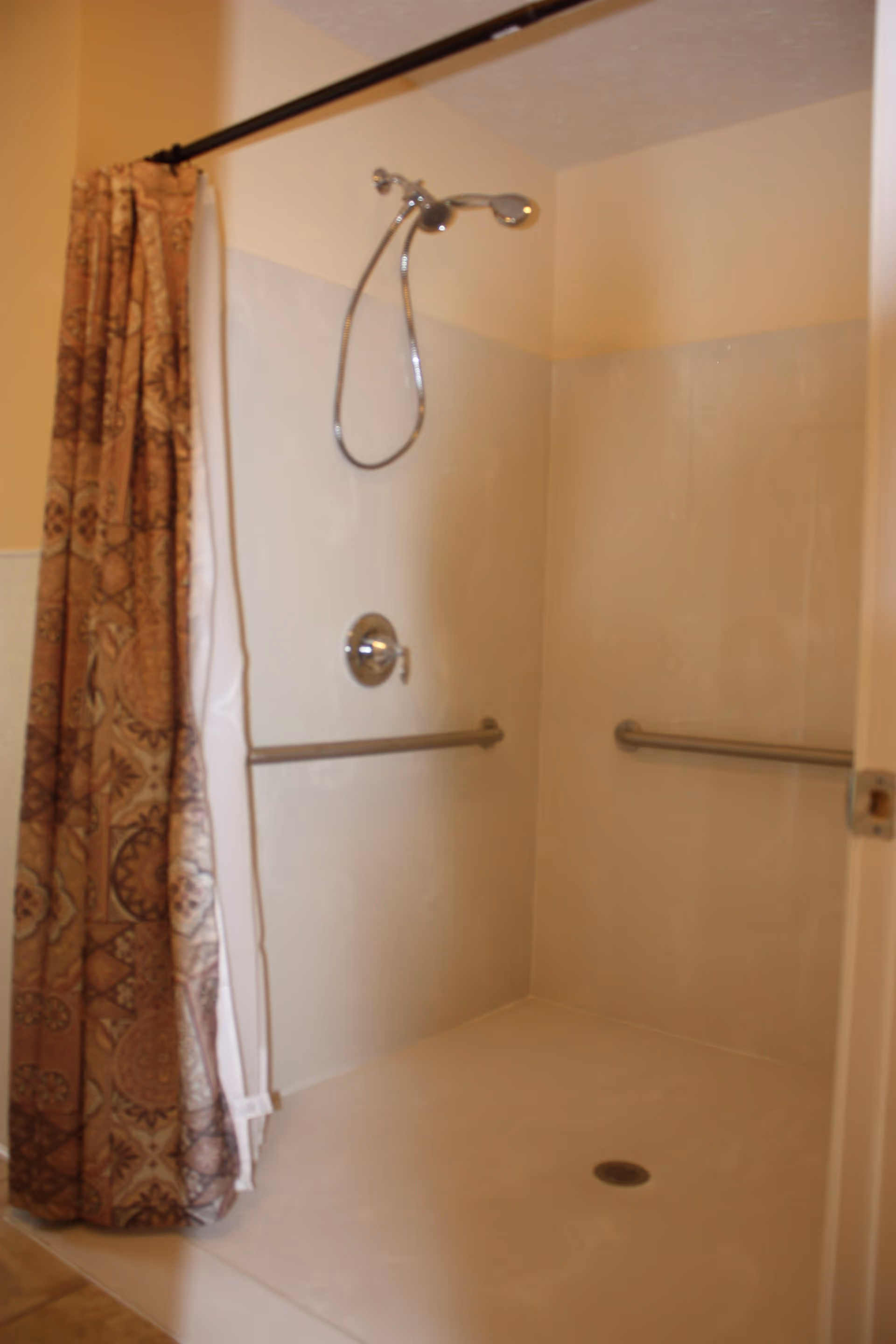 A walk-in shower with beige walls and floor, featuring two horizontal grab bars on the walls, a handheld showerhead attached to a flexible hose, and a patterned brown shower curtain partially pulled to one side.