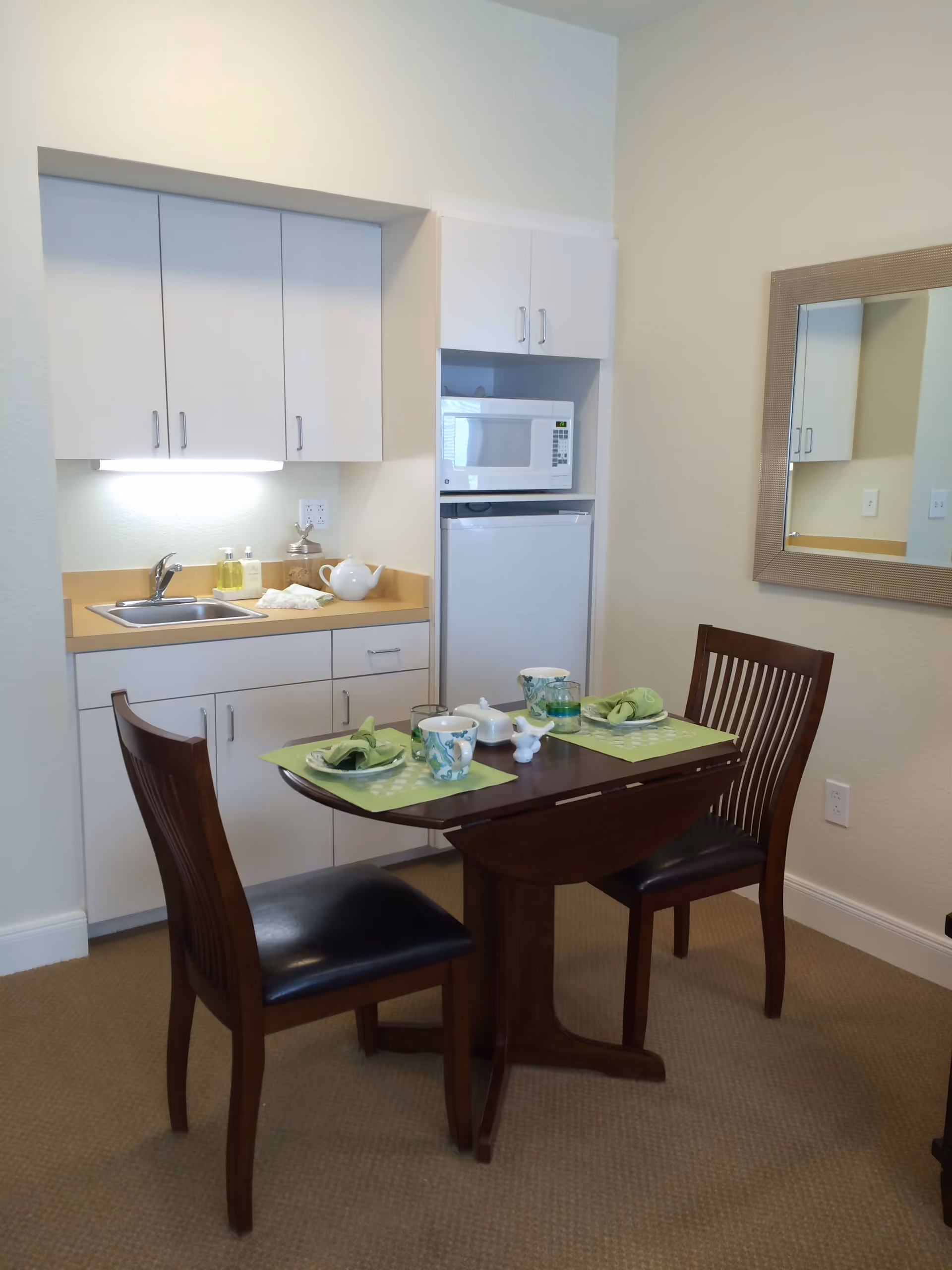 Small kitchenette area with white cabinets, a sink, a microwave, and a mini refrigerator. In front of the kitchenette is a dark wooden table set for two with green placemats, cups, and napkins. A large rectangular mirror hangs on the wall to the right.