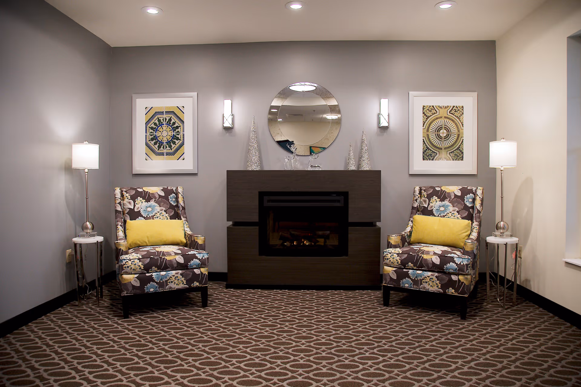 A cozy sitting area with two floral patterned armchairs each with a yellow cushion, positioned on either side of a modern dark wood fireplace. Above the fireplace is a round mirror flanked by two wall sconces. Two framed abstract artworks hang on the wall behind the chairs. Each chair is accompanied by a small round side table with a lamp. The room has a patterned carpet and neutral-colored walls.