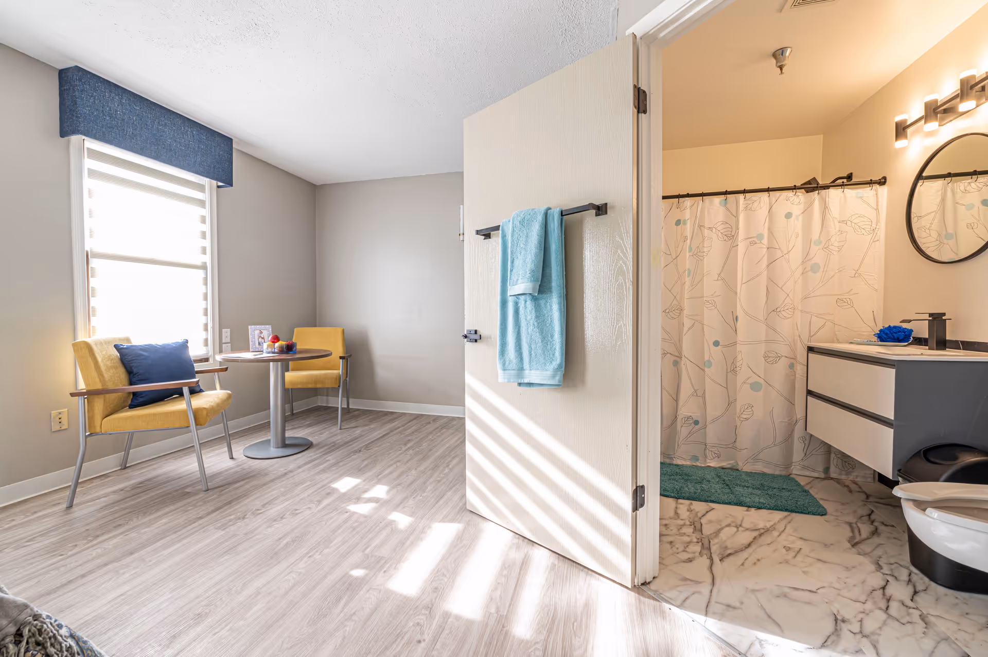 A bright room with light wood flooring featuring two yellow chairs with blue cushions around a small round table near a window with a blue valance. An open door reveals a bathroom with marble-patterned floor tiles, a shower curtain with a leaf pattern, a vanity with a round mirror, and a blue bath mat.
