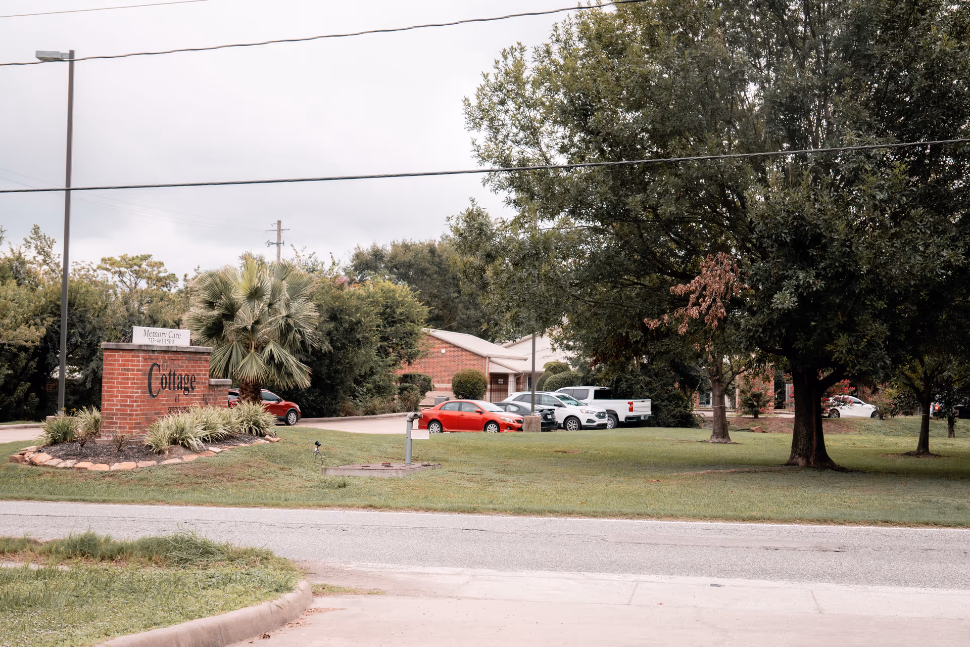 Exterior view of The Cottage of Spring Branch facility showing a brick sign with landscaping in front, several parked cars, and trees on a grassy lawn under an overcast sky.