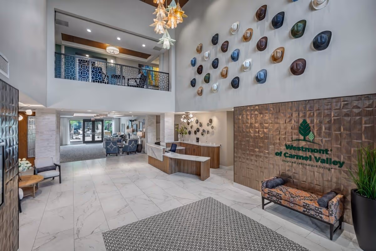 Spacious and modern lobby area of Westmont of Carmel Valley featuring a high ceiling with decorative hanging lights, a wall adorned with various hats, a reception desk, seating area with chairs and tables, and a large sign displaying the facility's name and logo.