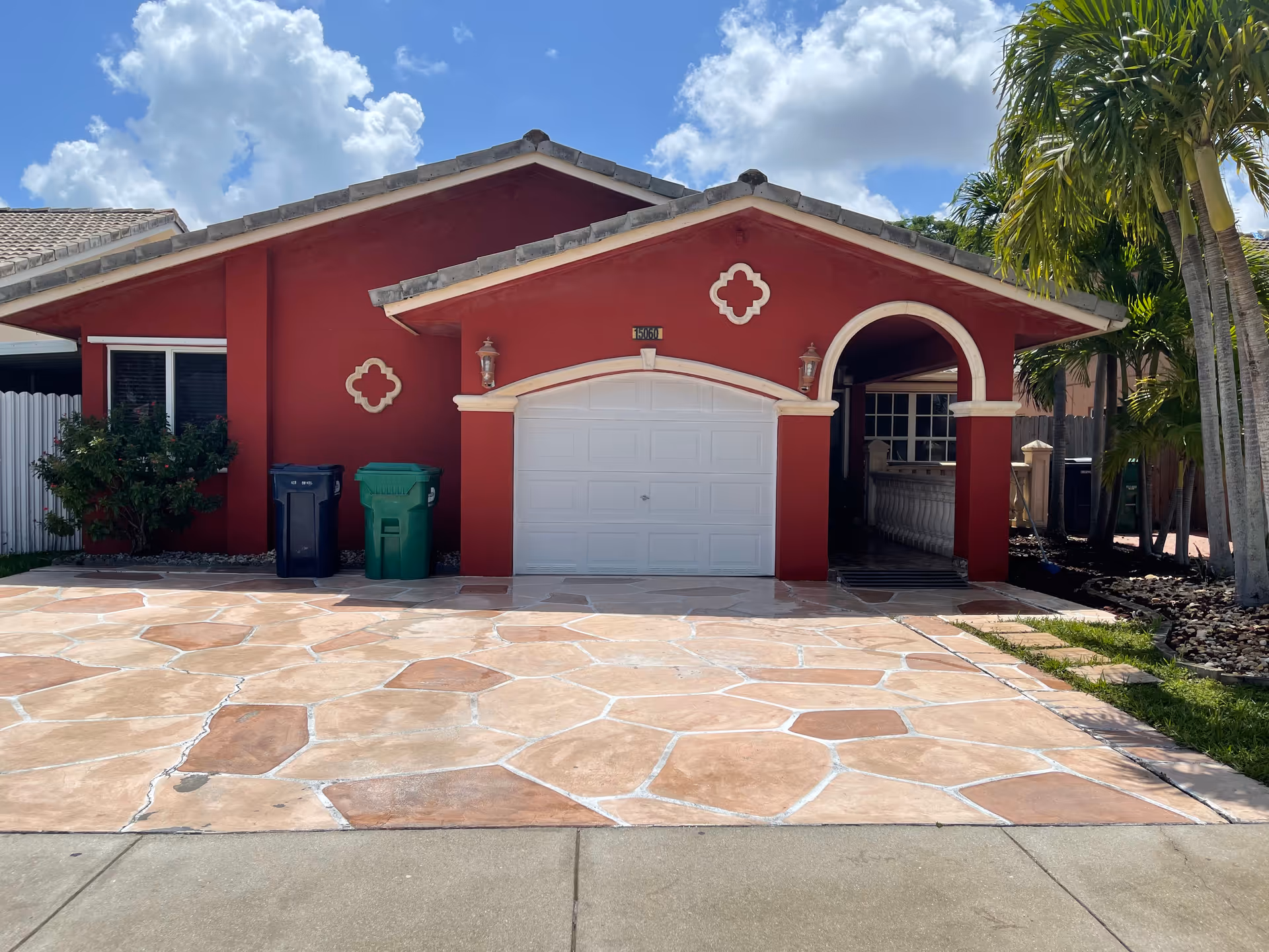 Front exterior view of a single-story red house with a white garage door, two trash bins on the left side, a tiled driveway, and palm trees on the right side under a partly cloudy sky.