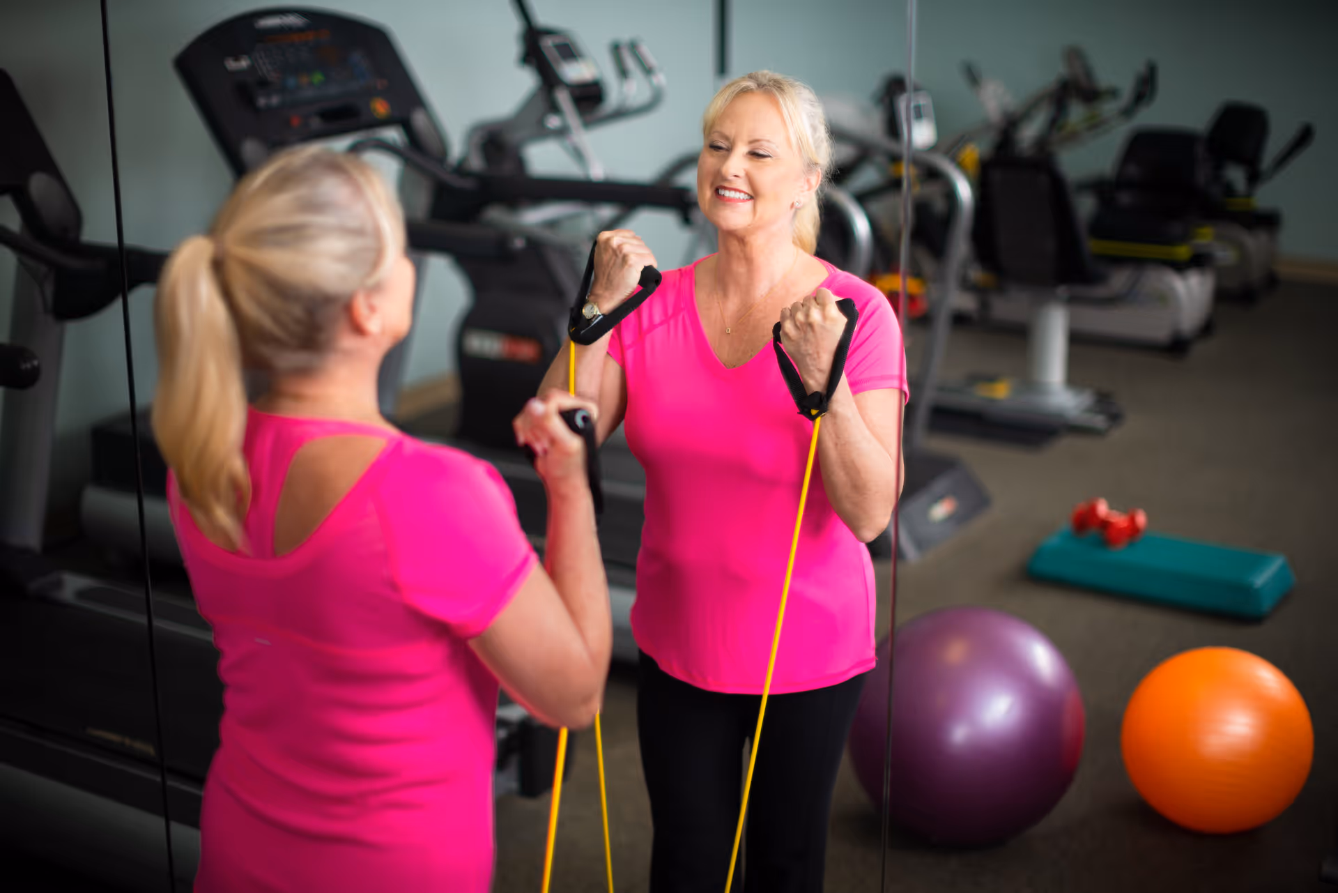 An older woman in a bright pink shirt exercises with resistance bands in front of a mirror in a fitness room. Behind her, there are various exercise equipment including treadmills, exercise balls, and weights.