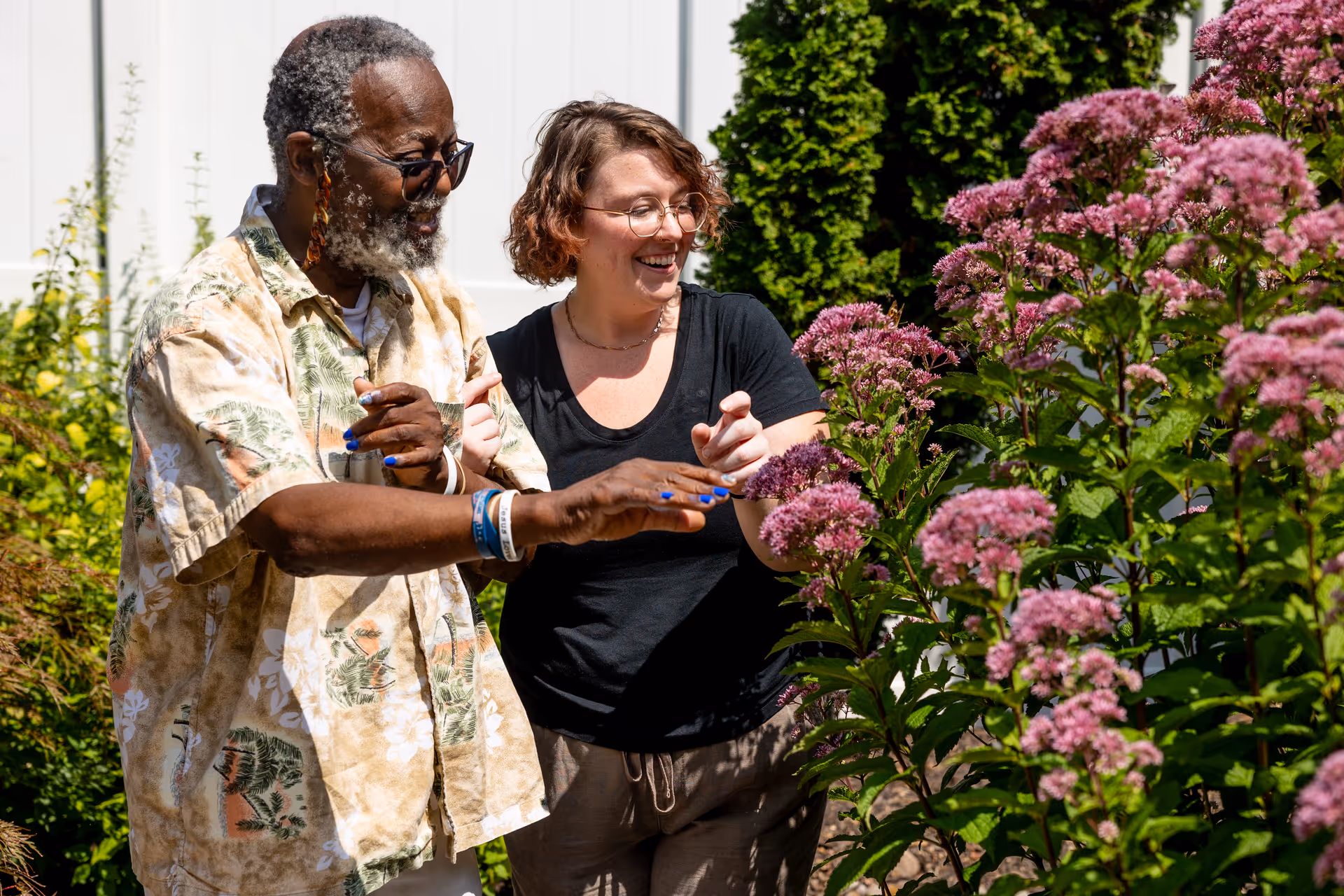 An elderly man and a younger woman are standing outdoors near a garden with tall pink flowers. The man is wearing a patterned short-sleeve shirt and sunglasses, while the woman is wearing glasses and a black shirt. They are both smiling and appear to be enjoying the flowers together.
