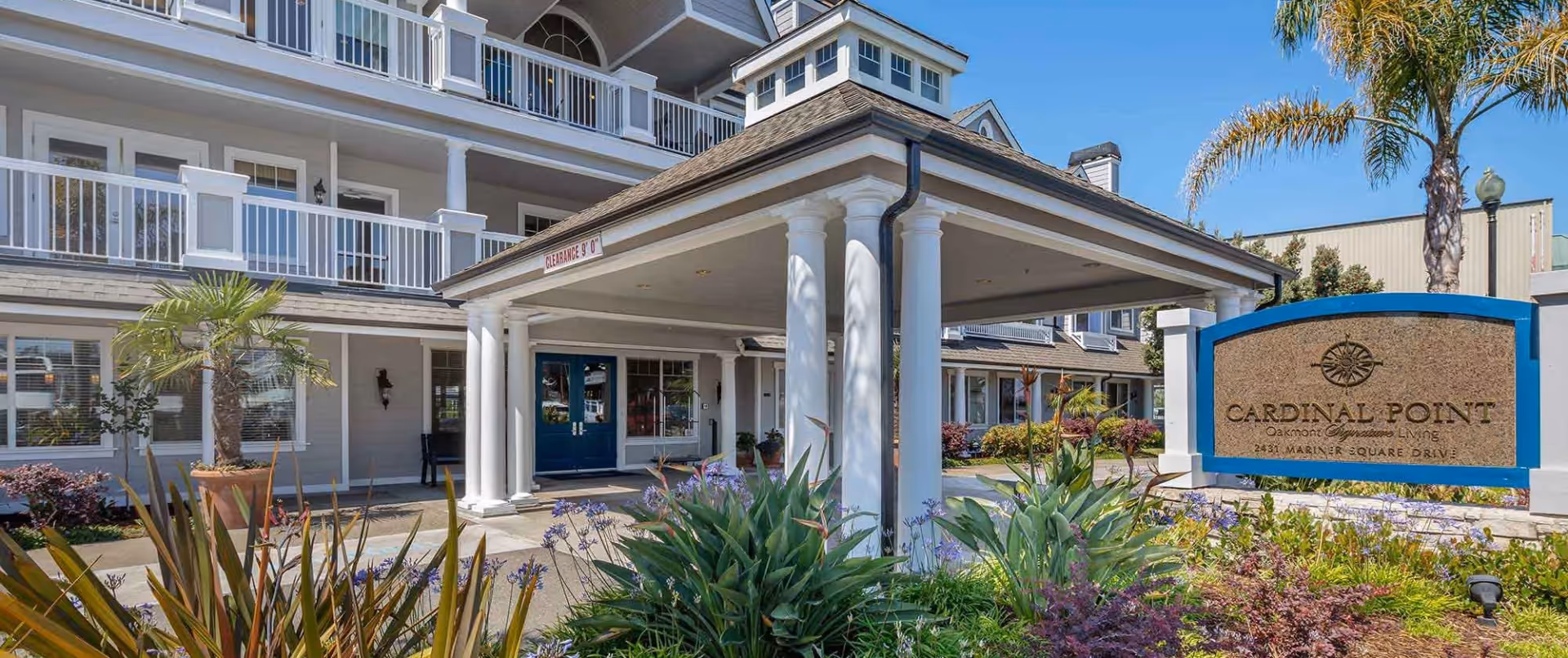 Exterior view of Cardinal Point senior living facility showing the entrance with white columns and a covered driveway. The building has multiple balconies and windows, surrounded by landscaped plants and palm trees under a clear blue sky.