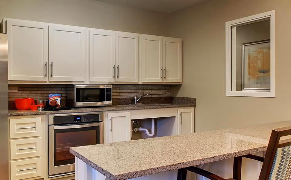 A modern kitchen area with white cabinets, a microwave, an oven, and a granite countertop island with chairs. There is a window on the right wall and some kitchen items like a red bowl and a boxed cake mix on the counter.