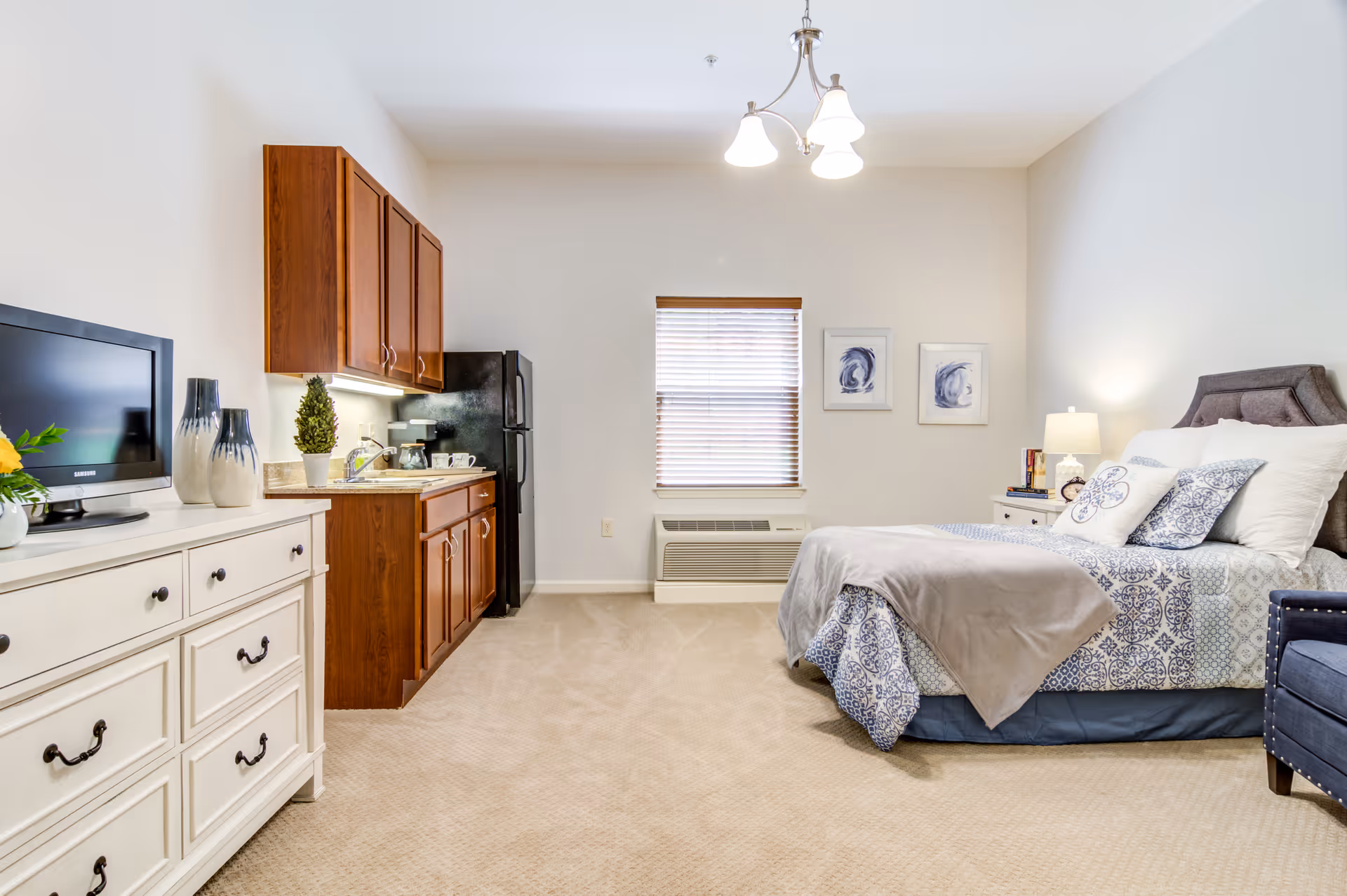 A bright and clean senior living studio apartment featuring a bed with patterned bedding and pillows, a nightstand with a lamp and books, a small kitchenette with wooden cabinets, a black refrigerator, and a countertop with a coffee maker. A white dresser with a TV and decorative vases is on the left side. The room has beige carpet, a window with blinds, and two framed abstract artworks on the wall.