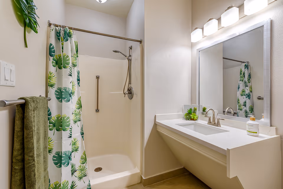 A clean and modern bathroom featuring a walk-in shower with a green and white leafy patterned shower curtain, a green towel hanging on a towel rack, a white countertop with a sink, a large mirror above the sink, and three light fixtures mounted above the mirror. There is also a small decorative plant on the countertop.