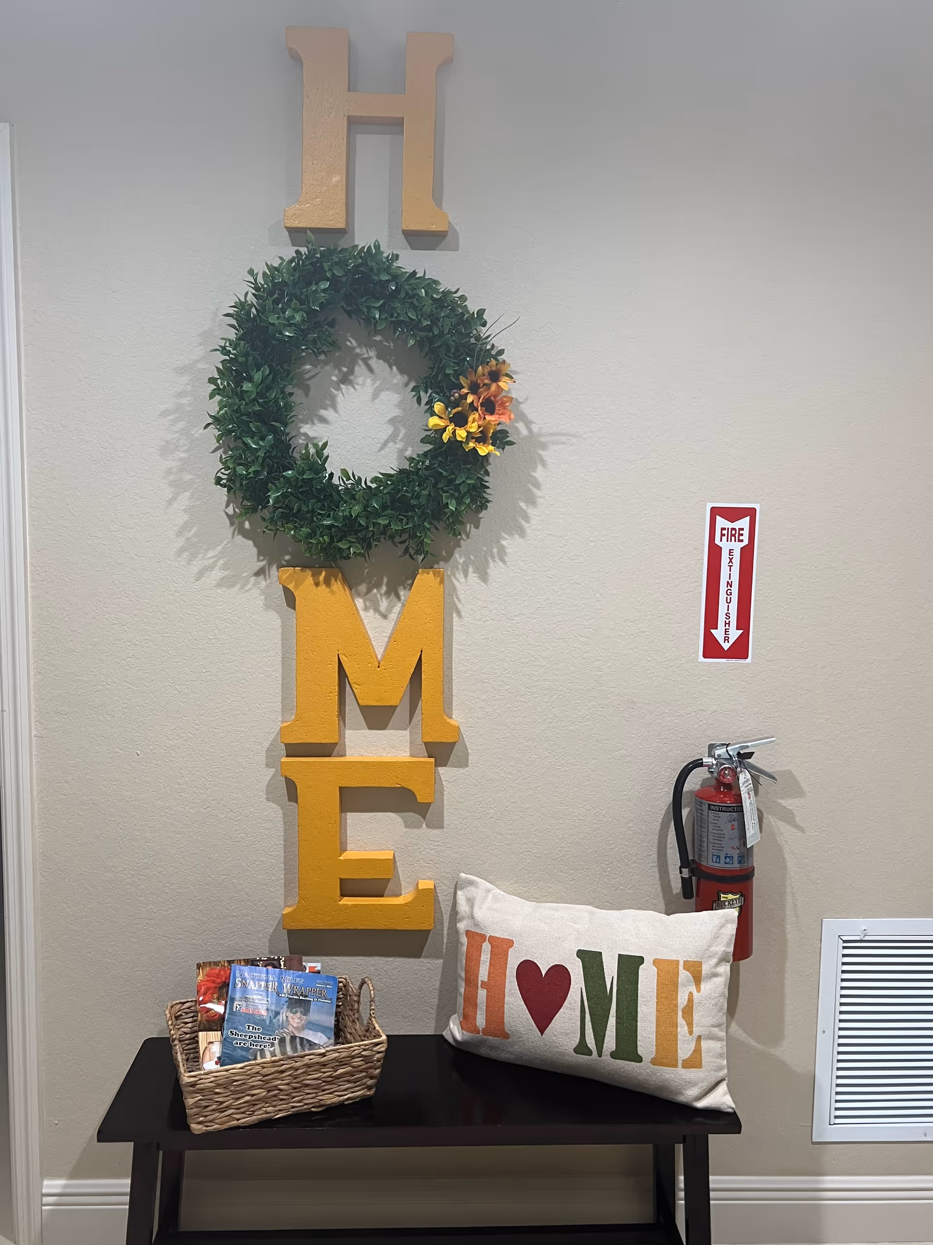 A decorative wall display with large yellow letters spelling 'HOME' vertically, with the letter O replaced by a green wreath adorned with small flowers. Below the letters is a black bench holding a basket with magazines and a pillow with the word 'HOME' printed in colorful letters with a red heart replacing the letter O. A fire extinguisher is mounted on the wall to the right.