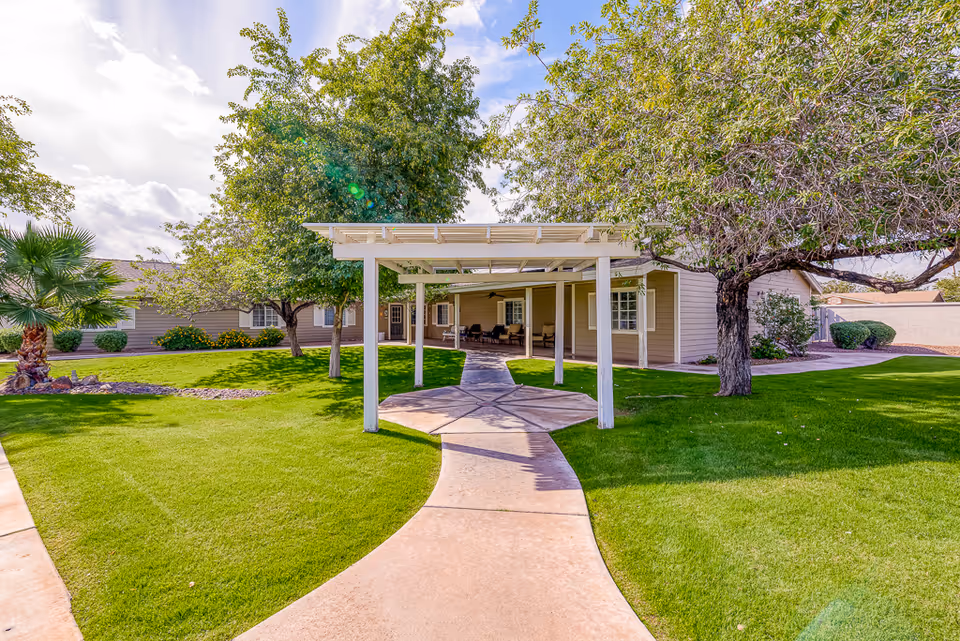 A well-maintained outdoor garden area at Paradise Valley Senior Living featuring a curved concrete walkway leading to a covered pergola. The area is surrounded by green grass, trees, and shrubs with a beige building in the background under a partly cloudy sky.