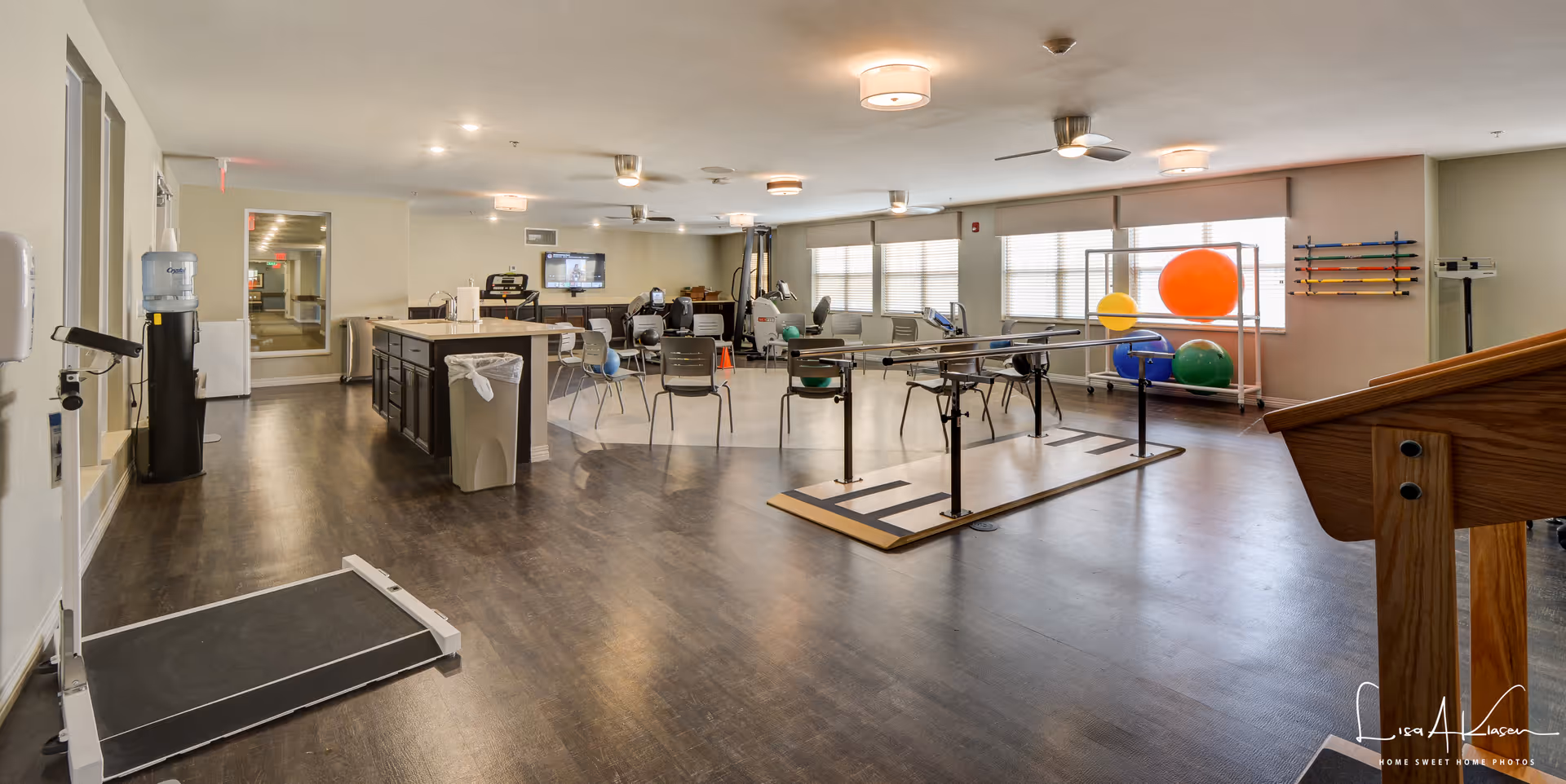 A spacious rehabilitation or physical therapy room in a senior living facility with exercise equipment including parallel bars, therapy balls, chairs, and a treadmill. The room has large windows allowing natural light, wood flooring, and neutral-colored walls. There is a water dispenser and a counter with a trash bin, and a TV mounted on the far wall.