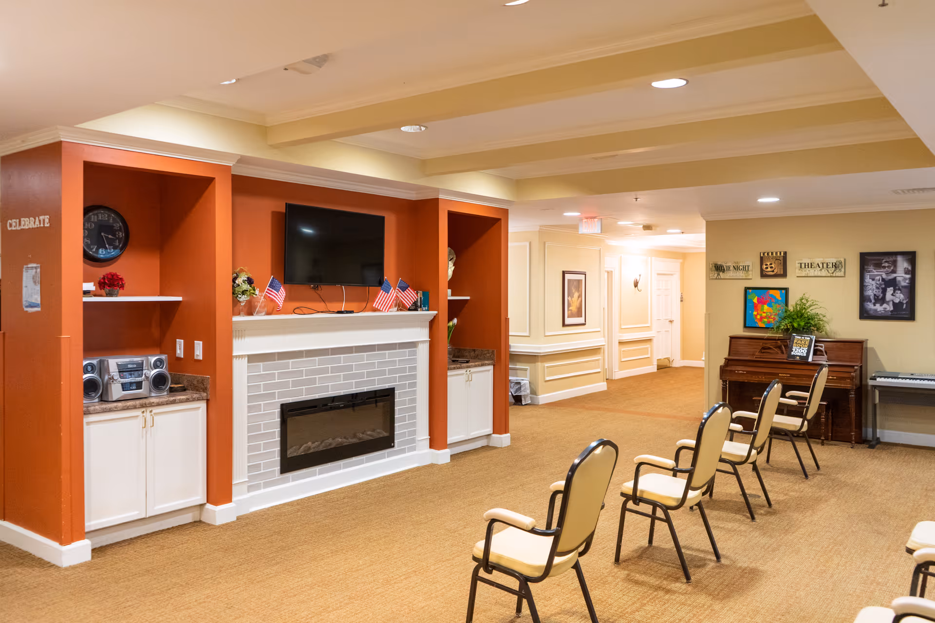 Interior view of a senior living facility common area with beige carpet and walls. There is a white brick fireplace with a mounted flat-screen TV above it, flanked by orange accent walls with built-in shelves and cabinets. Several beige chairs with armrests are arranged in rows facing the fireplace. On the right side, there is a piano with a plant and framed pictures on the wall above it. The ceiling has recessed lighting and beams.