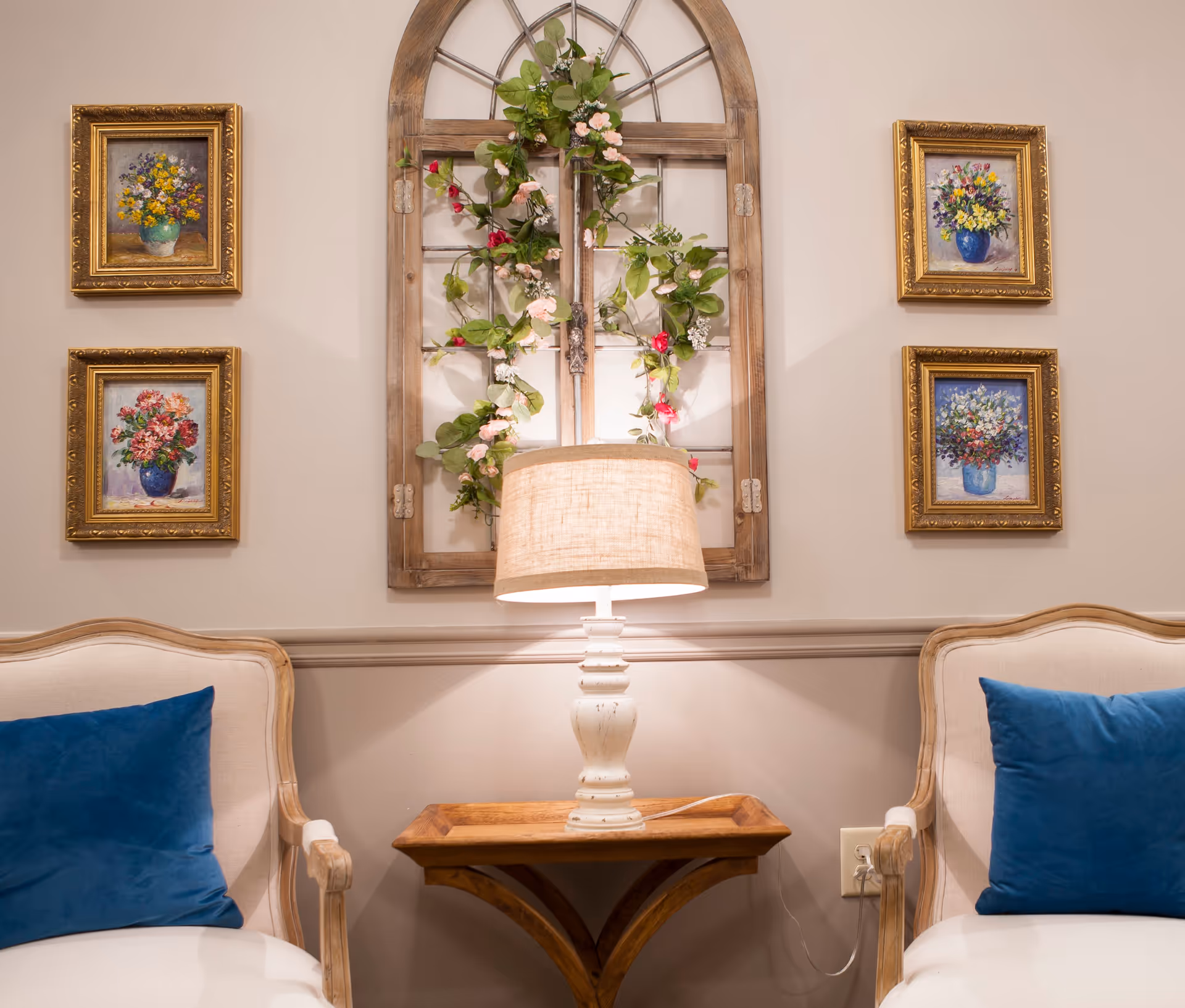 A cozy interior seating area with two beige armchairs featuring blue cushions, a wooden side table with a white table lamp, and a decorative wooden window frame adorned with green and pink floral garlands. Four framed floral paintings hang on the wall, two on each side of the window frame.