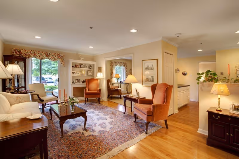 A cozy living room with wooden floors, a large patterned area rug, two orange upholstered armchairs, a cream-colored sofa, a wooden coffee table with candles and a small plant, and a side table with a lamp. There are built-in shelves with decorative items and a window with floral curtains letting in natural light. The room opens into another area with a desk and chair, and there are framed pictures on the walls.